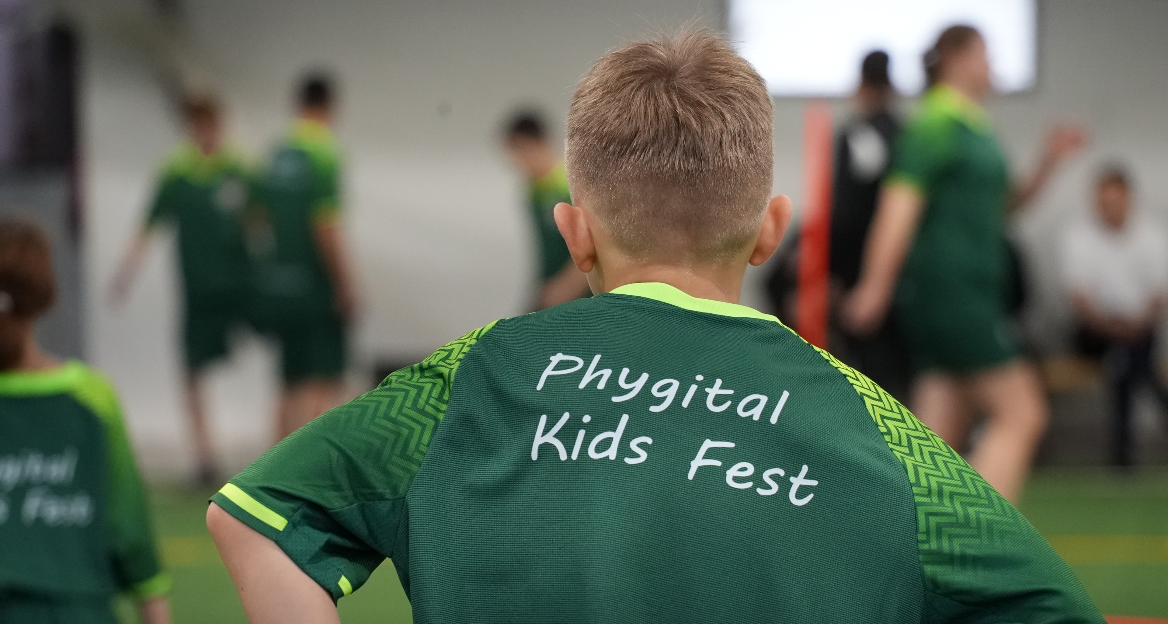 Артынан түсірілген бала жасыл футболкада, арқасында Phygital Kids Fest жазуы, манежде