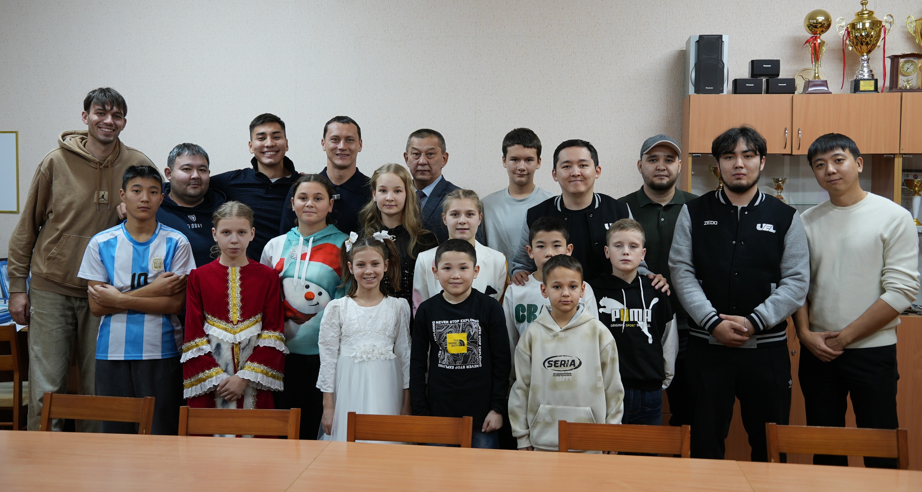 Балалар мен QOFA өкілдері акт залында топтық суретте, сөреде кубоктар мен марапаттар