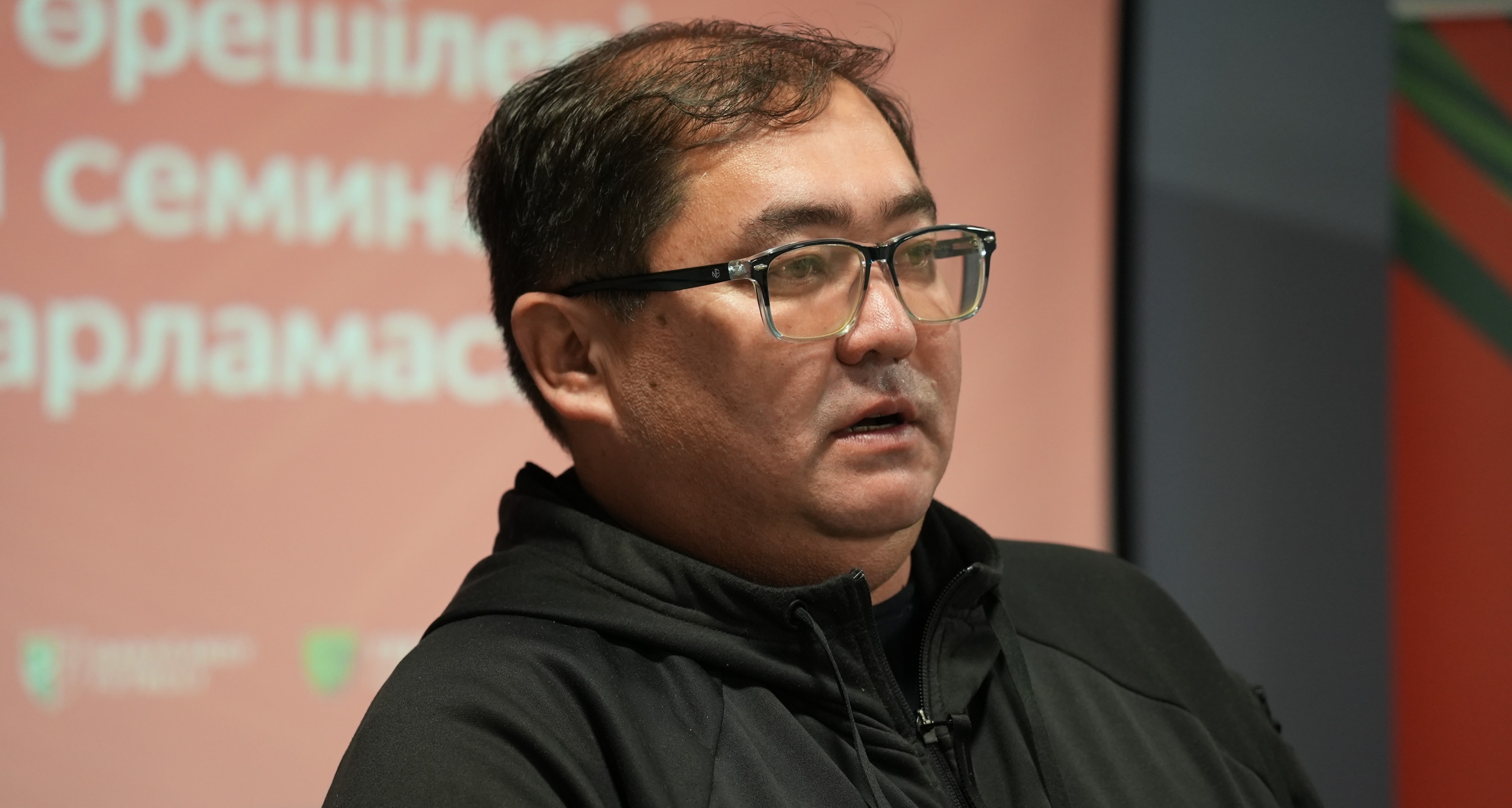 Мейрам Серіков семинарда сөз сөйлеуде, артында QOFA презентациясының экраны